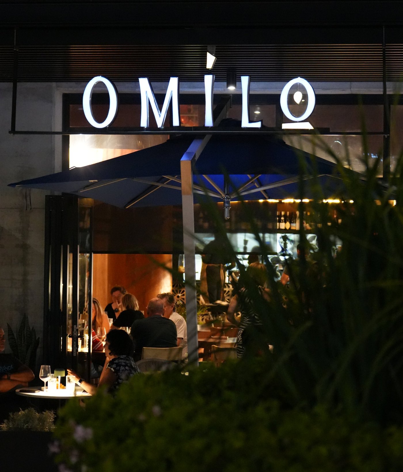 The Omilo shopfront at night, the lettered sign lit above a full dining room